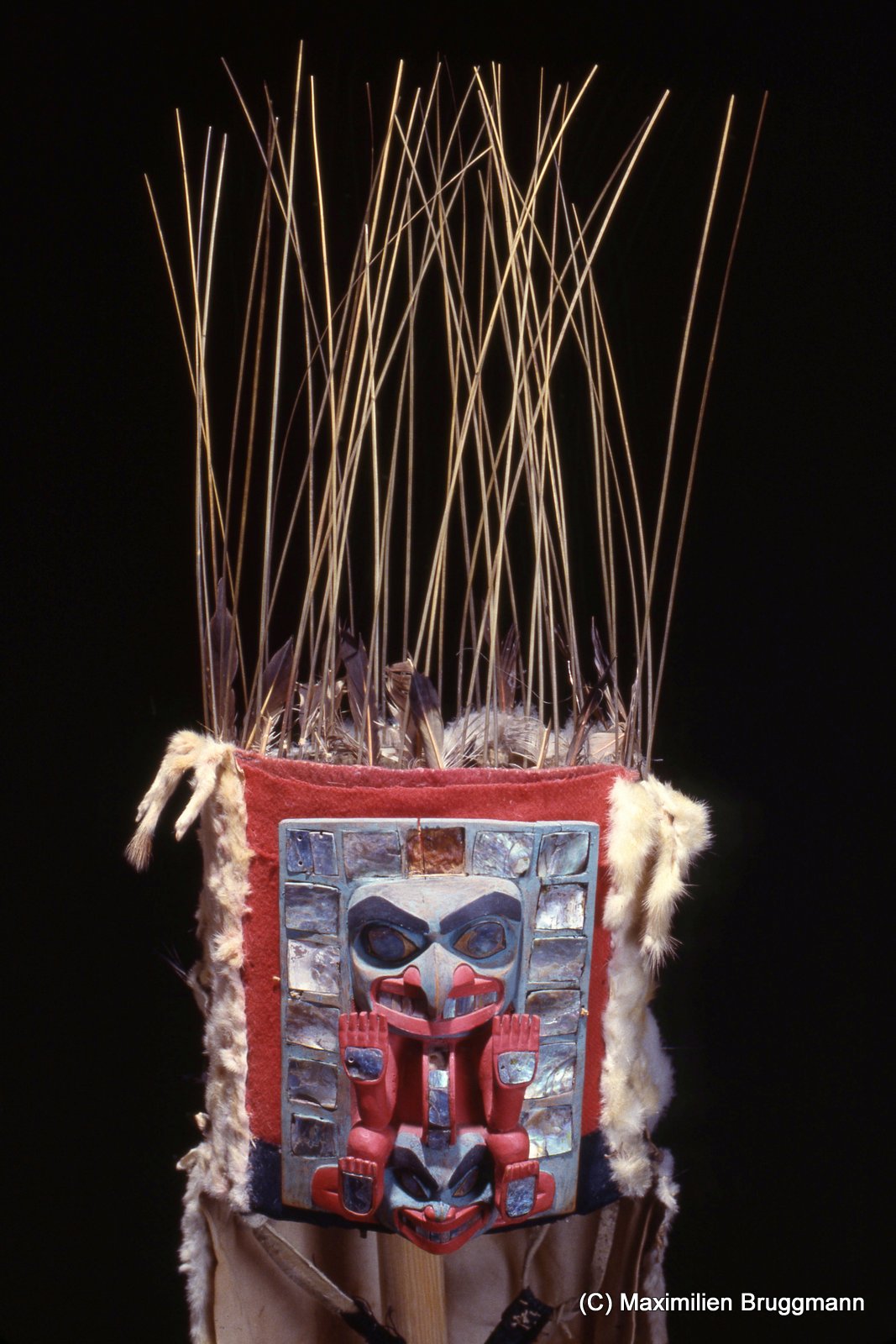 14 Especially impressive as examples of the art of mask making are the crown=like frontlets of the northern coastal people. A chief would wear one of these decorative headpieces during the dance to welcome his guests. The mask shown depicts what is presumably an eagle, below which is a human face. The mask is made of hard wood, haliotis, sea lion whiskers and eagle down; "fringe" is of ermine skins. (Mask 19 cm, extender 85 cm; SJM)